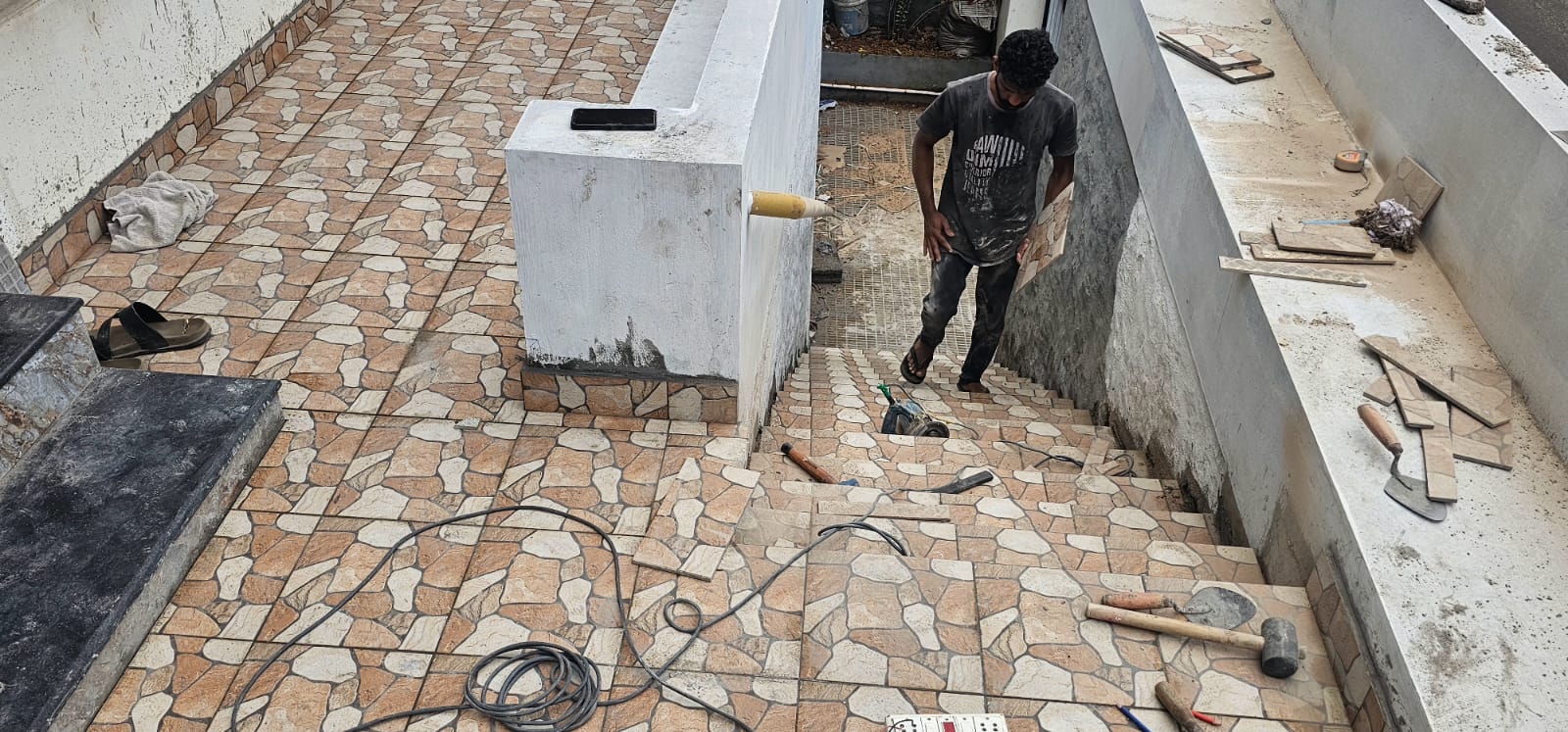 Skilled construction worker installing natural cobblestone-pattern floor tiles on an outdoor terrace at an AKS Constructions project site, with trowels and tile tools visible.